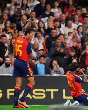 A soccer match photo showing two Spanish national team players, with player #15 standing and player #19 (Lamine Yamal) kneeling on the ground while fans watch from the stands.