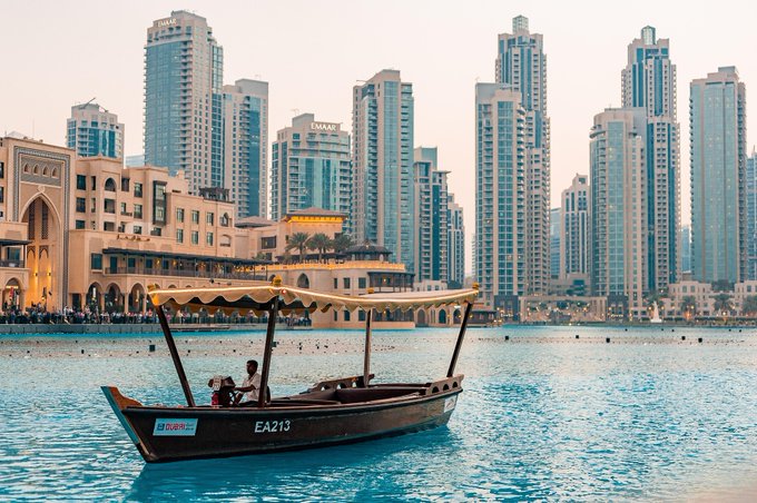 A traditional abra boat marked "EA213" carries passengers across a Dubai waterway, set against a backdrop of towering skyscrapers and waterfront architecture. The serene yet disciplined journey across the water mirrors the post's message of steady progress and unwavering grind toward long-term goals.