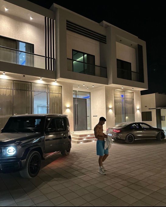 A shirtless man in blue shorts stands in a driveway between a gray Mercedes G-Wagon and a sedan, in front of a modern illuminated house at night.