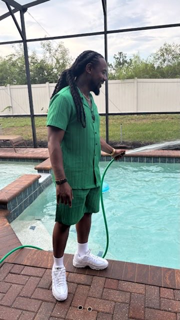 A man with long dreadlocks wearing a matching green shirt and shorts set stands beside a swimming pool, holding a green garden hose while smiling. The image visually echoes the post's "liquidity" and "green candles" wordplay, using the green hose and pool as a creative metaphor for market optimism.