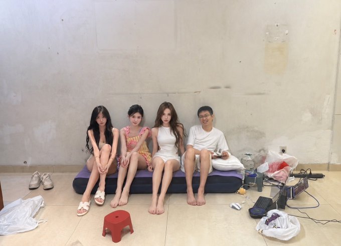 Three young women and one young man sitting together on a small couch against a plain concrete wall, surrounded by various items on the floor.