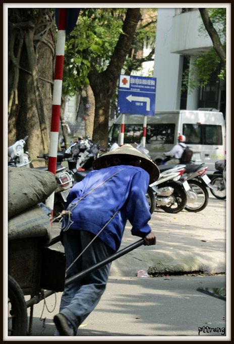 A street vendor wearing a conical hat and blue jacket pulls a cart down a street lined with parked motorbikes and trees in Vietnam, with a directional sign visible in the background.
