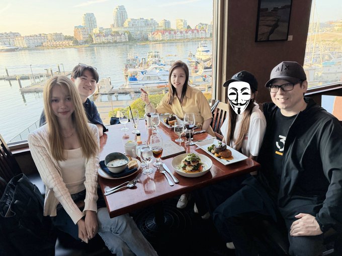 Five people gather around a restaurant table with plates of food and wine glasses, enjoying a meal with a waterfront marina and city skyline visible through the large windows behind them, appearing to celebrate a birthday for @Excellion.