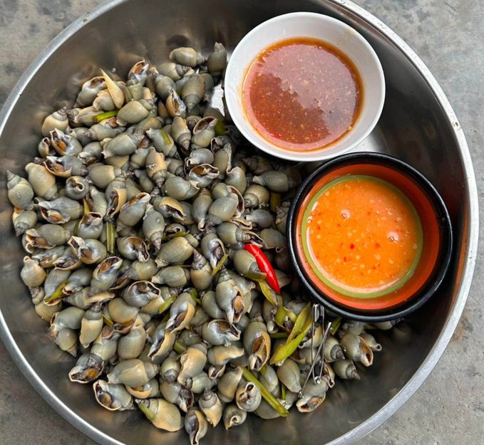 A metal bowl filled with cooked snails is served with two dipping sauces in small bowls, garnished with red chili peppers and lemongrass.