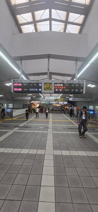 Interior view of a Japanese train station with a clock and electronic departure boards showing times like 15:01 and 15:43, with travelers walking through the tiled concourse.
