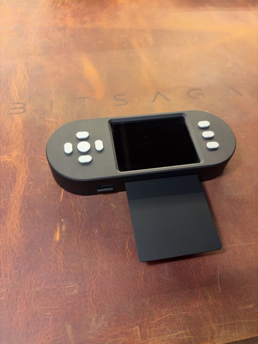 A handheld gaming device with a screen and white control buttons resting on a wooden surface with the engraved text "B I T S A G K", featuring a black stand/attachment on its back.