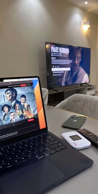 A laptop displays the Netflix page for the series "Peaky Blinders" on a table, with a TV screen in the background also showing the show's title and information, suggesting someone unwinding with the series after a long day.