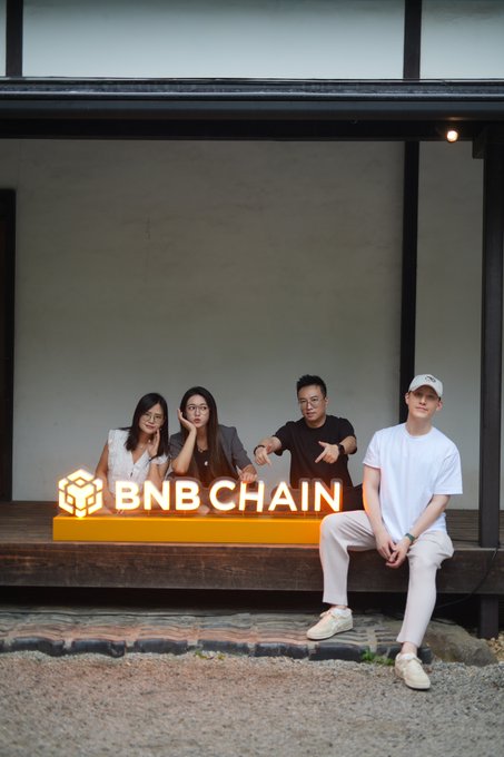 Four people posing with illuminated BNB CHAIN signage on a wooden bench outside a modern building.