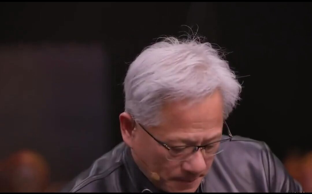 The image shows an older man with white hair wearing glasses, sitting at a desk or table while looking down at something. The background appears to be a dark room, which contrasts with the man's appearance. There are other people present in the scene but they are not the main focus of the image.