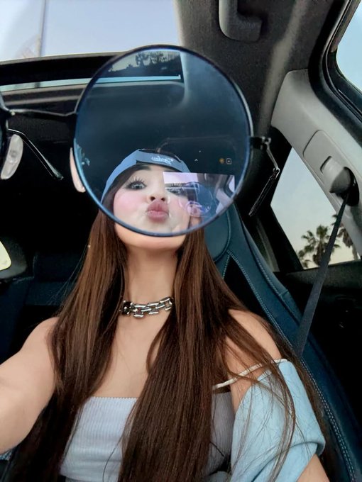A young woman takes a selfie in a car, making a kissing face reflected in the rearview mirror, wearing a silver chain choker and a light blue top.