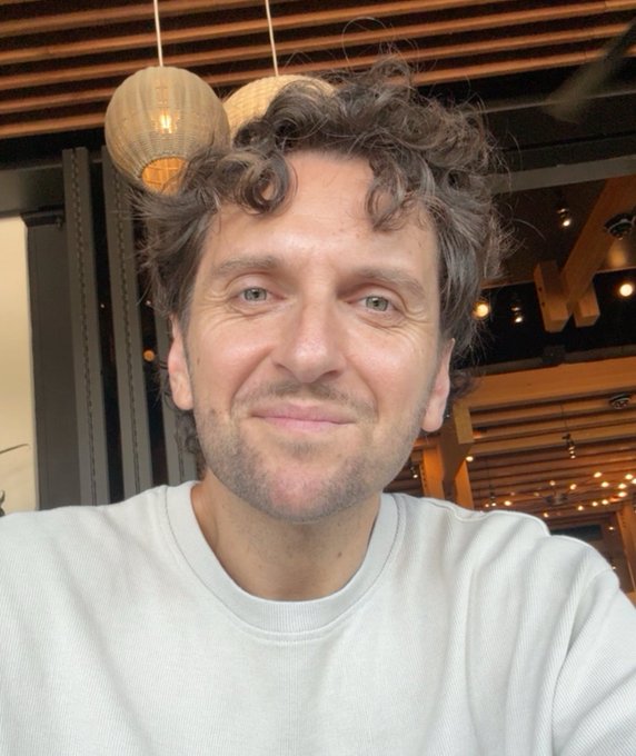 A selfie of a smiling man with curly hair wearing a white shirt in what appears to be a restaurant with hanging lights.