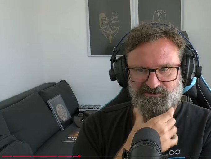 A bearded man with glasses and headphones sits in front of a microphone in a room with a dark couch and framed artwork on the wall behind him.