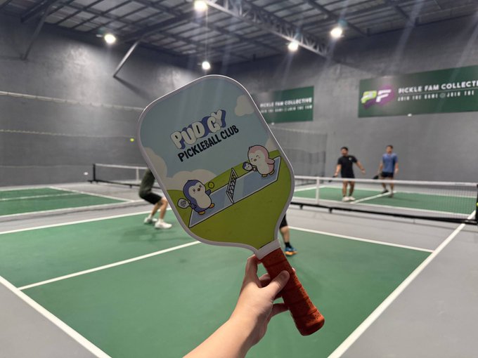 A first-person view of someone holding a PUCK Pickleball Club branded paddle at an indoor pickleball facility, with other players visible on the court in the background.