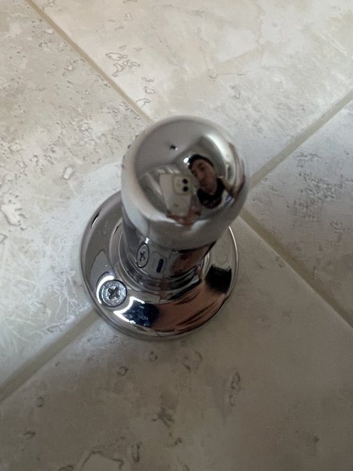 A close-up photo of a shiny chrome doorknob reflecting a person holding a smartphone, mounted on a beige tiled surface.