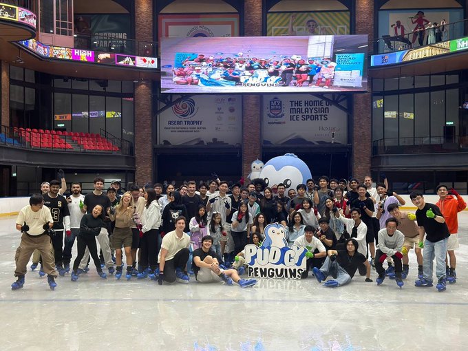 A group photo of ice skaters posing on an ice rink in front of a 'Pudgy Penguins' mascot and signage for 'The Home of Malaysian Ice Sports'.