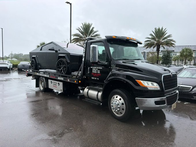 A black flatbed tow truck, operated by "9 Brothers Towing" (visible on the door along with a phone number), hauls a matte black stealth military-style armored vehicle. The post jokingly questions whether this massive, armored-looking purchase was funded by crypto money, highlighting the vehicle's unique, almost covert appearance.