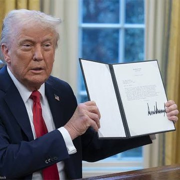 The image shows a man wearing a suit, sitting at a desk with an open letter or bill in front of him. The text on the document appears to be about Donald Trump's signature. The background features a window that allows natural light into the room.