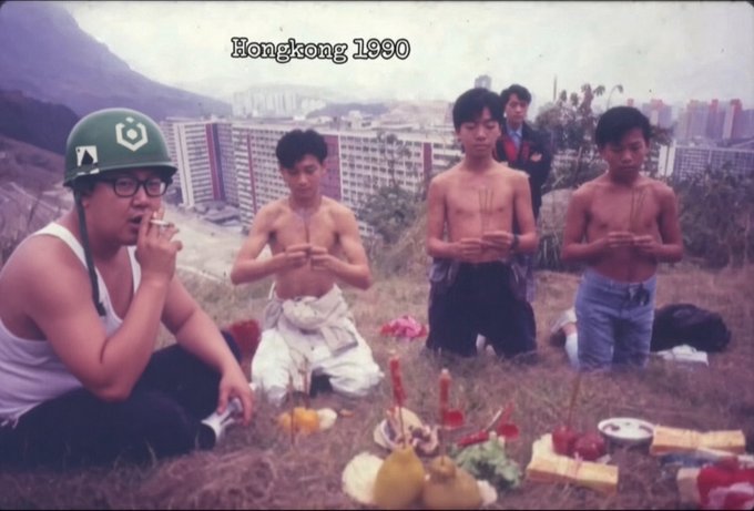 A vintage photograph labeled 'Hongkong 1990' showing a group of shirtless young men and a man in a helmet making offerings with incense at what appears to be an outdoor ritual site with a city skyline in the background.