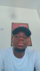 A selfie of a man wearing a black cap and white t-shirt, with a framed artwork hanging on a beige wall behind him.