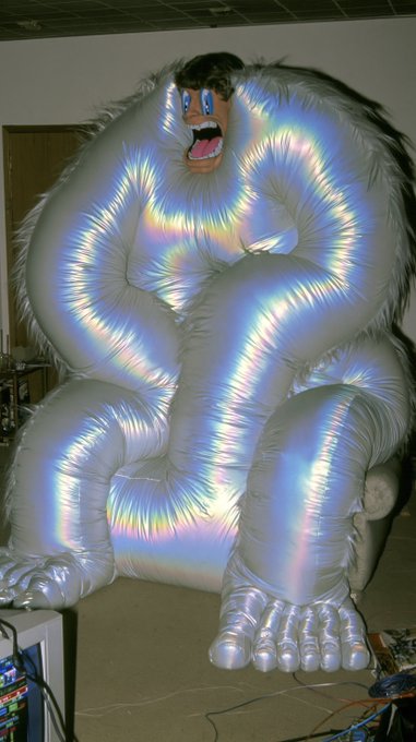 A large inflatable gorilla figure illuminated with iridescent rainbow lighting sits in what appears to be a storage or work area with a mask for a face.