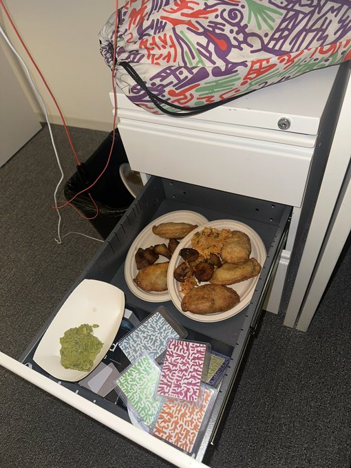 Photo of an open office desk drawer containing two plates of empanadas or fried pastries, a bowl of green guacamole, and some colorful notebooks, with a graffiti-style backpack resting on top of the desk.