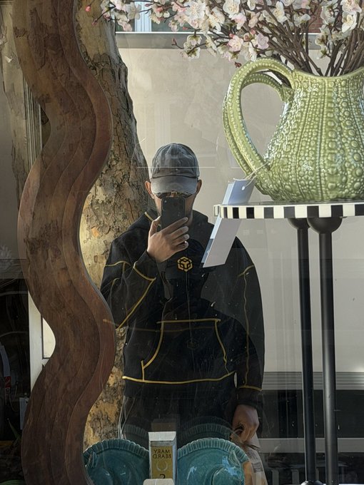 A mirror selfie of a person wearing a black cap, sunglasses, and dark jacket, standing in what appears to be a store with a large green decorative pitcher and floral arrangements visible.