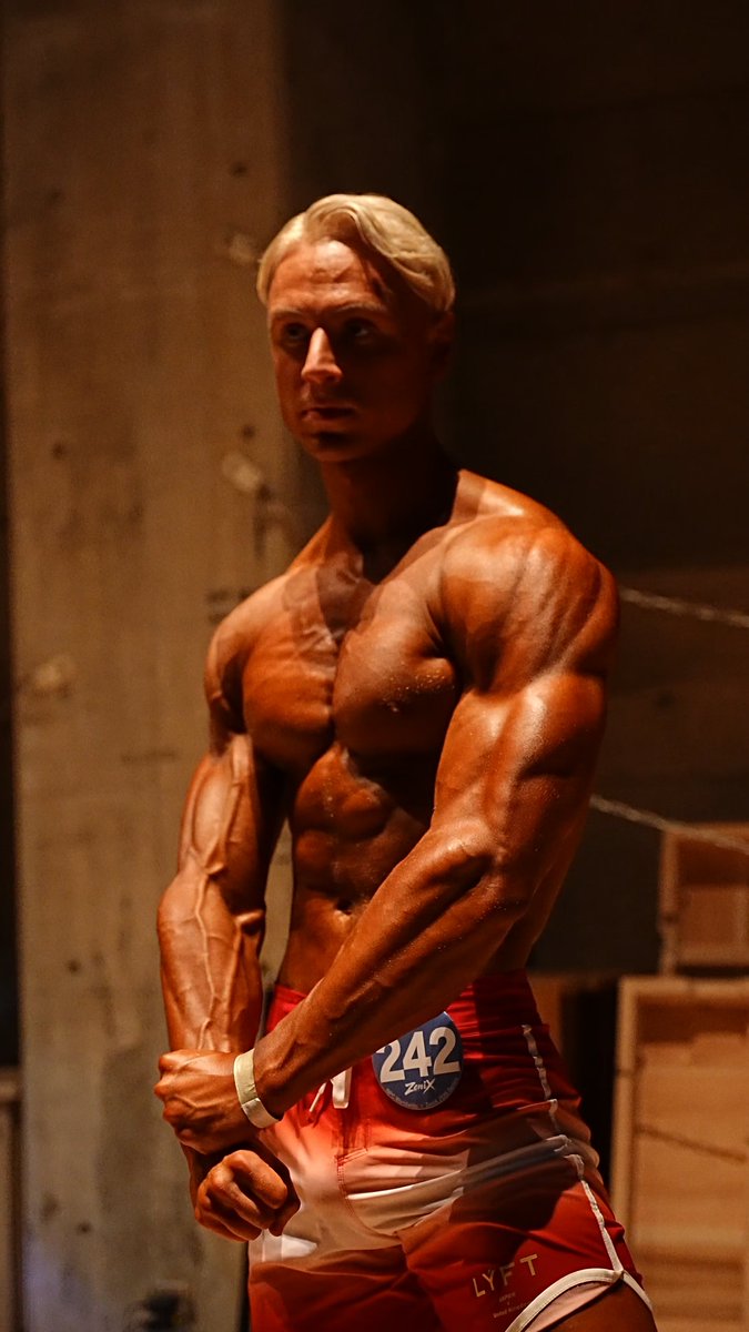 A blonde, heavily-muscled bodybuilder with a bronzed tan poses on stage wearing red LYFT shorts and competitor number "242," captured right before stepping out for his pre-pump performance.