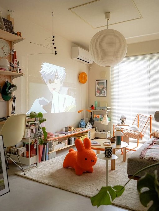 A cozy, well-decorated room with a paper lantern, house plants, and an orange bunny-shaped chair, featuring an anime character being projected onto the wall.