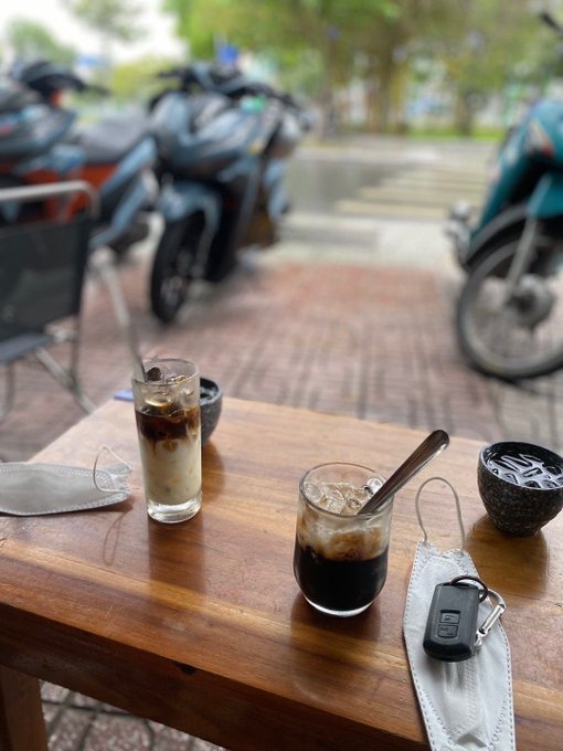 A wooden outdoor cafe table holds two glasses of iced coffee, a small bowl with spoons, a face mask, and a Honda car key, with motorcycles parked in the blurred background.