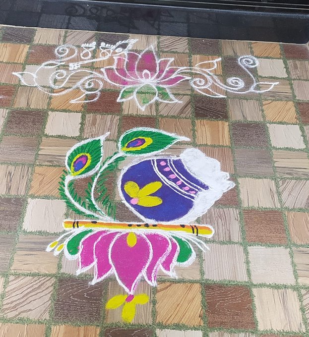 A vibrant Rangoli artwork displayed on a tiled floor features a peacock feather, a pot, a flute, and a lotus flower, designed with colorful powders.