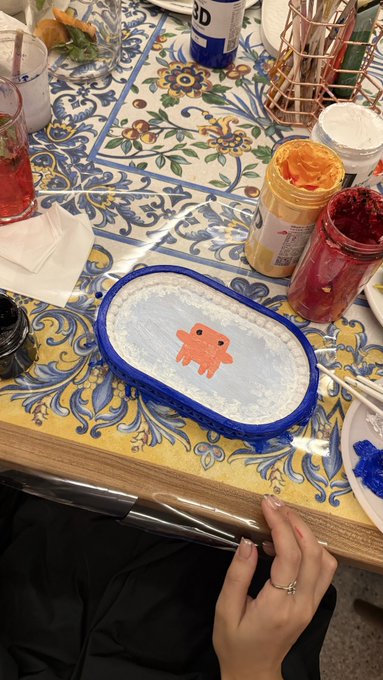 A hand wearing a ring rests on a decorative tiled table next to a blue and white painted dish featuring a cartoon character, with jars of paint nearby.