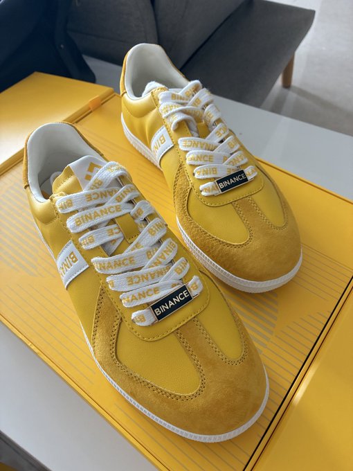 A pair of yellow sneakers with white laces printed with 'BINANCE' branding, displayed on a yellow shoebox.