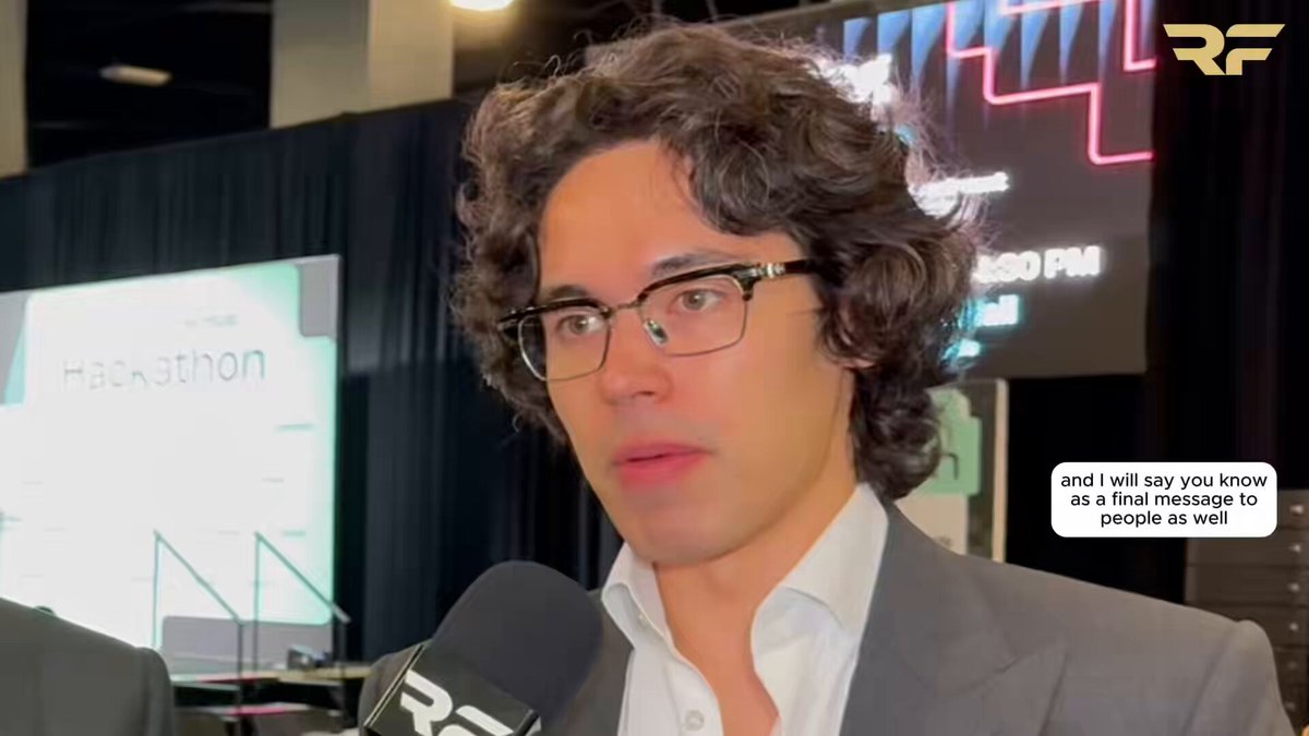 The image features a man wearing glasses, standing behind a microphone at an event or conference. He appears to be giving a speech or interview, as indicated by his position near the camera. The background of the image shows a stage with a banner that reads "Hackathon", suggesting that this is likely a tech-related gathering or competition.