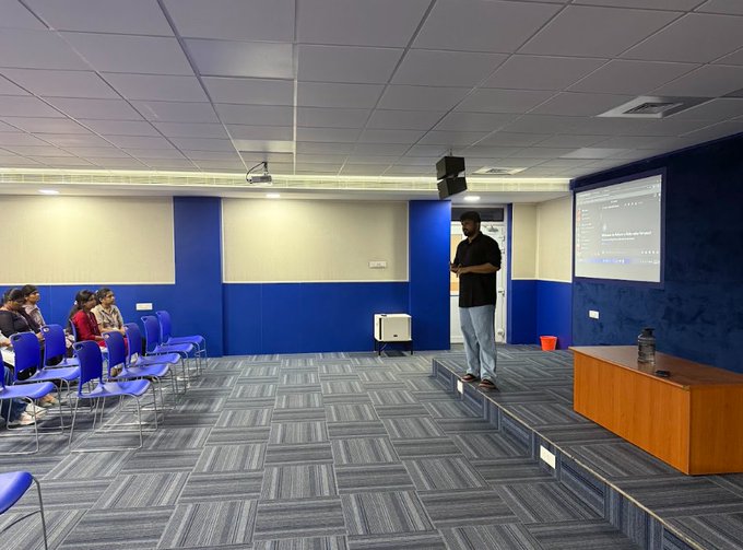 A man stands on a stage in a presentation room with blue walls and a projection screen, facing a small seated audience in a conference setting.