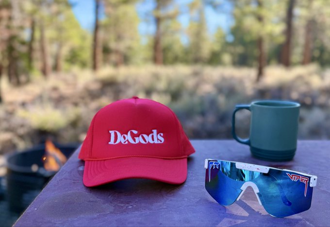 A red DeGods baseball cap and a pair of TFRST sunglasses rest on a table next to a green ceramic mug, with a campfire and pine trees visible in the background.