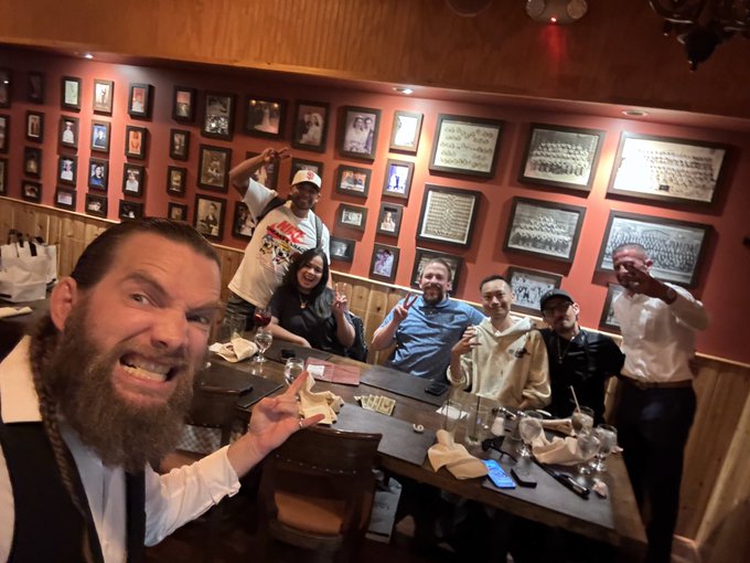 A group selfie taken at a restaurant with a bearded man in the foreground, surrounded by friends posing at a dining table with framed photos covering the walls behind them.