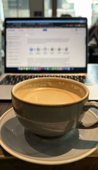 A ceramic cup of coffee sits on a saucer in the foreground, with a laptop screen displaying a webpage in the blurred background.