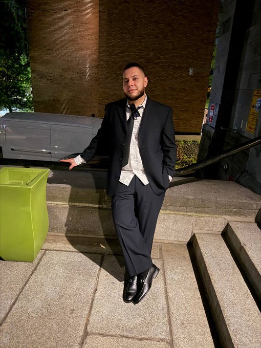A young man in a black tuxedo with bow tie leaning against a green planter on stone steps at night.