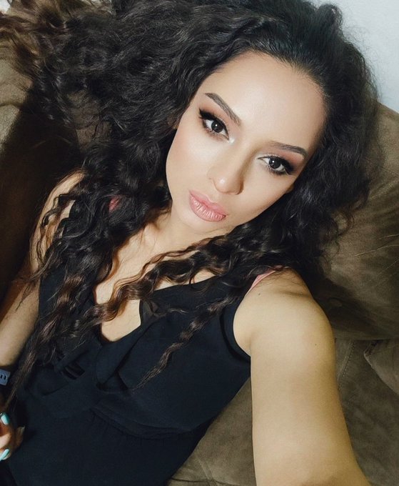 A young woman with long, voluminous curly black hair poses for a selfie wearing a black ruffled top.