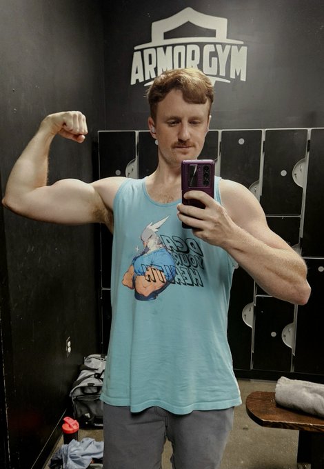A man takes a mirror selfie flexing his bicep at Armor Gym while wearing a light blue tank top featuring an anime-style graphic design.