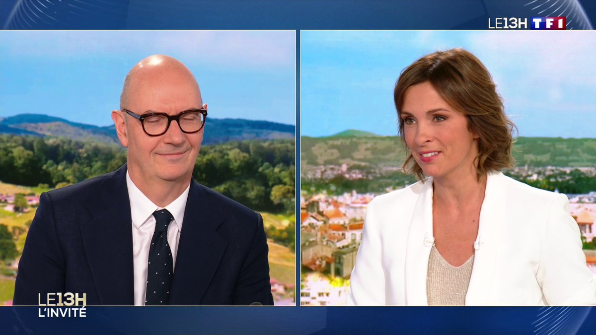 A screenshot from the French TV program 'LE 13H' on TF1 showing two news anchors—a bald man with glasses and a woman in a white blazer—with the label 'L'INVITÉ' at the bottom.