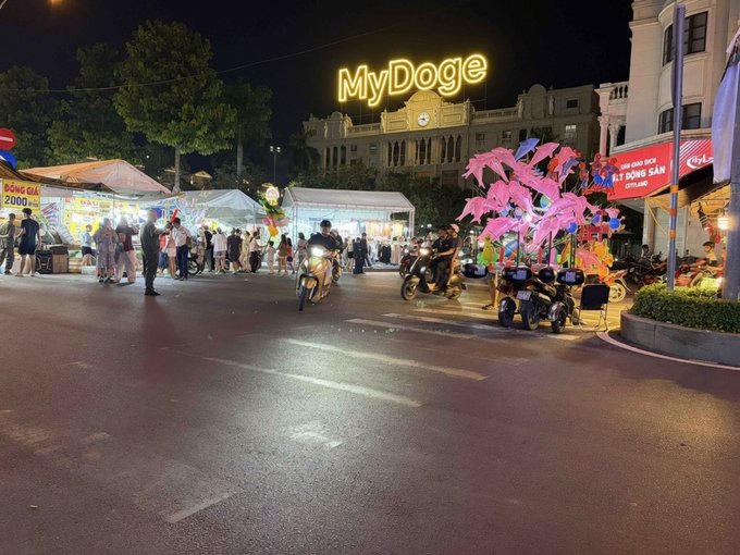 A nighttime street scene in Vietnam with a large illuminated 'MyDoge' neon sign, a vendor selling 'Bông Gòn 2000', and decorative pink lantern trees lining the road with motorcyclists passing by.
