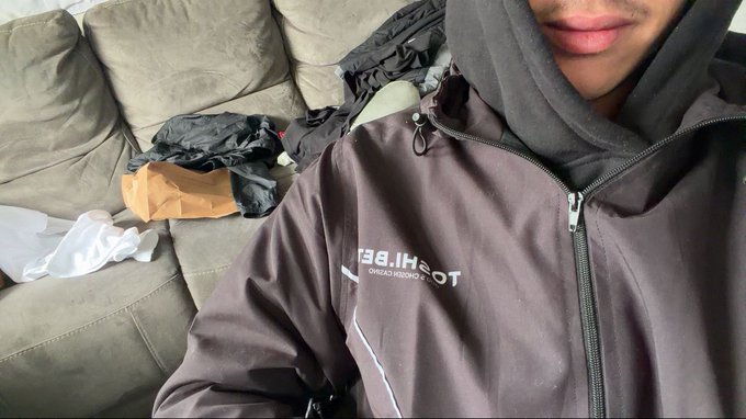 A person wearing a dark gray jacket with "TOSHI B.F.T. Tokyo Design Studio" text visible on the chest, paired with a hood, while reclining on a gray couch with scattered items behind them. The image showcases their new Toshi tech apparel, aligning with the post celebrating the brand.
