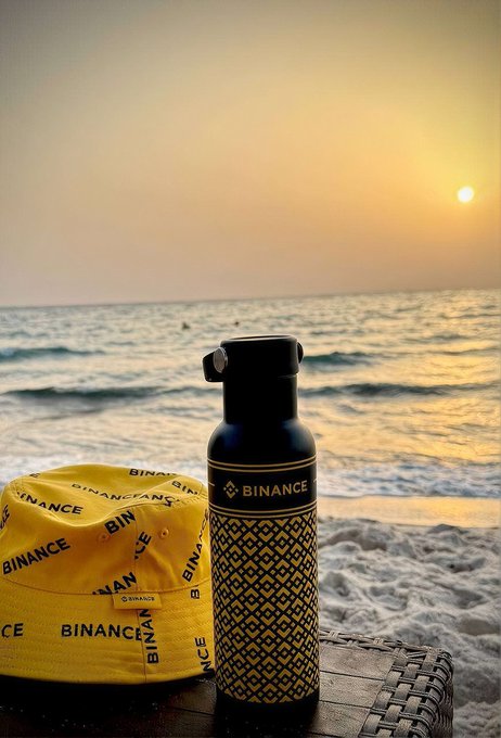 A yellow "BINANCE" branded cap and a sleek black water bottle sit on a wicker table by the ocean at sunset, capturing the perfect crypto-meets-coastal vibe for a sunny escape.