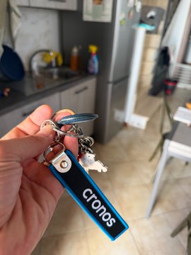 A hand holds a keychain with keys and a blue tag reading 'cronos', with a kitchen visible in the background.