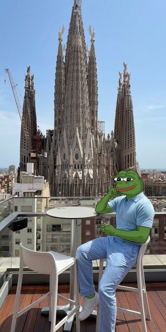 A Pepe the Frog character sitting at an outdoor café table with a drink, overlooking the Sagrada Família basilica in Barcelona.