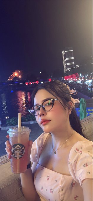 A selfie of a young woman with glasses holding an iced Starbucks drink at night, with a bridge and fireworks visible in the background.
