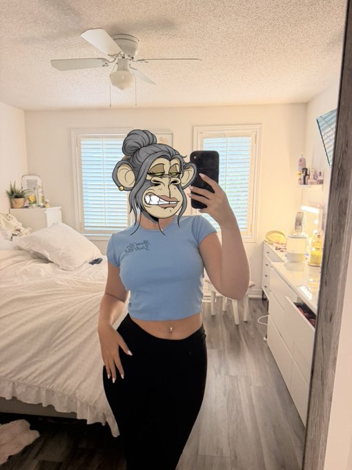 Bedroom selfie of a person wearing a light blue cropped t-shirt and black pants, holding a phone, with a cartoon monkey filter covering their face, standing in a room with a ceiling fan, bed, dresser with toiletries, and windows with blinds.
