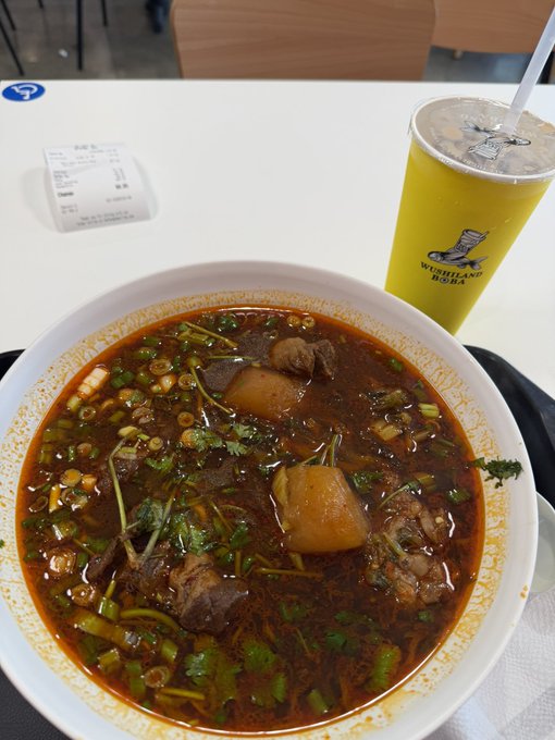A photograph of a bowl of spicy beef noodle soup with greens and a yellow Highland Soda drink on a white table with a receipt.
