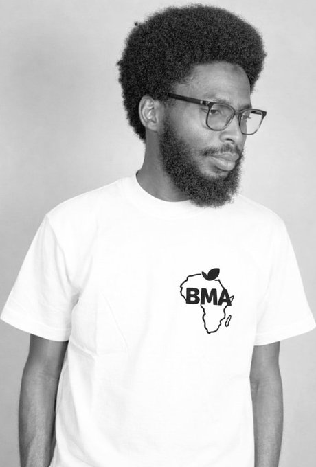 A black-and-white portrait of a man with a beard and glasses wearing a white t-shirt featuring an outline of Africa with 'BMA' written across it.